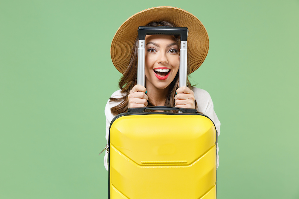 Close up satisfied fun traveler tourist woman in casual clothes hat hiding with yellow suitcase valise isolated on green background Passenger travel abroad weekend getaway Air flight journey concept